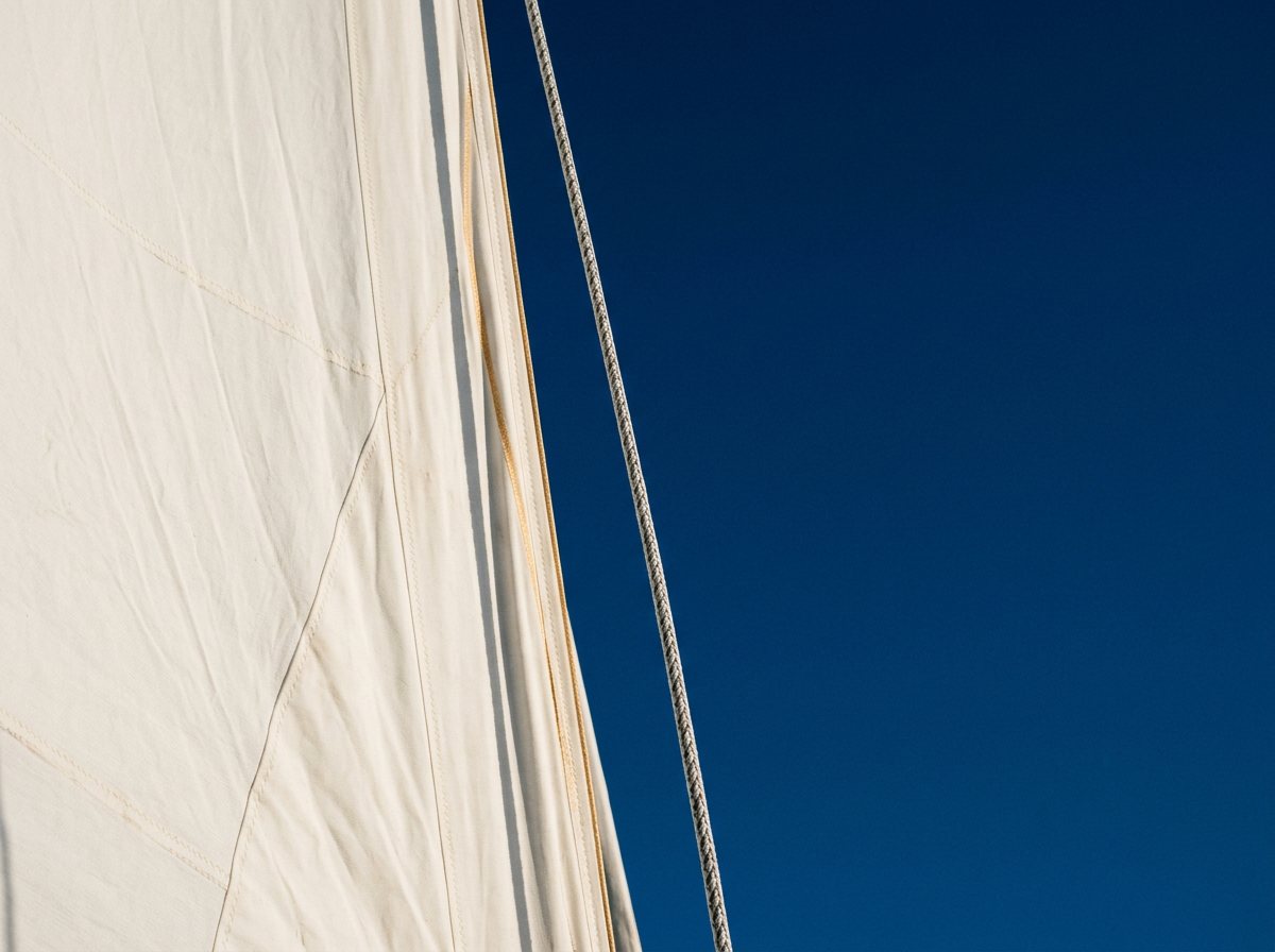 Detail eines weißen Segels vor tiefblauem Himmel, entlang der Segelkante ein feiner goldener Lichtstreif.
